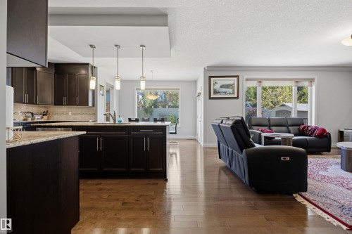 Open-concept living area featuring rich wood-finish flooring, dark wood cabinetry, a central kitchen island with seating, and ample natural light through large windows - 2043 Cameron Ravine Way Nw, Edmonton, AB - Indoor