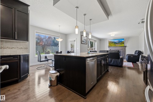 Open-concept kitchen and living area featuring wood-finish flooring, dark wood cabinetry with granite countertops, a central island with pendant lighting, and stainless steel appliances - 2043 Cameron Ravine Way Nw, Edmonton, AB - Indoor Photo Showing Kitchen With Upgraded Kitchen