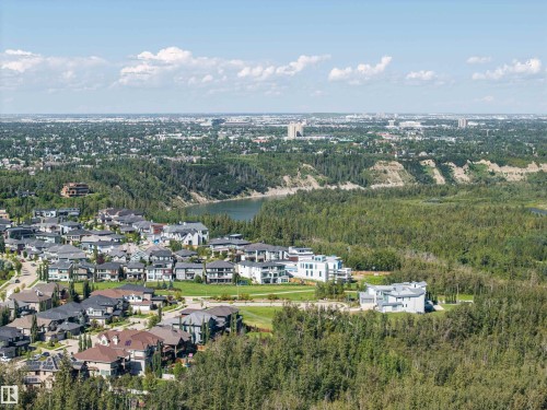 Aerial perspective of a residential development with varied architectural styles, surrounded by dense natural landscaping, a prominent body of water, and distant urban structures - 2043 Cameron Ravine Way Nw, Edmonton, AB - Outdoor With View