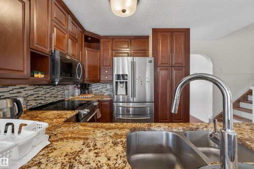 Kitchen featuring wood cabinetry, stainless steel appliances, a tile backsplash, and stone-finish countertops with an undermount double basin sink - 9007 169 Avenue Nw, Edmonton, AB - Indoor Photo Showing Kitchen With Double Sink With Upgraded Kitchen