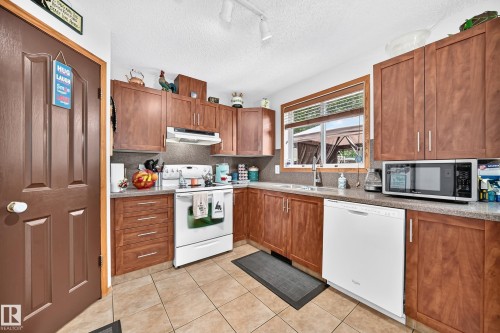 Kitchen featuring wood-finish cabinetry, tile flooring, solid surface countertops, and an integrated white dishwasher - 13069 37 Street Nw, Edmonton, AB - Indoor Photo Showing Kitchen