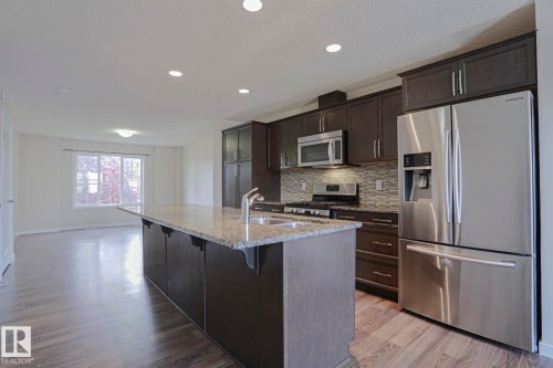 Kitchen featuring dark wood-finish cabinetry, stainless steel appliances, a tiled backsplash, and an island with a light-toned countertop - 308 Watt Boulevard Sw, Edmonton, AB - Indoor Photo Showing Kitchen With Upgraded Kitchen