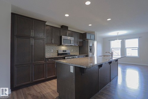 Kitchen featuring dark wood-finish cabinetry, stainless steel appliances, a tiled backsplash, a kitchen island with stone countertops, and wood-finish flooring - 308 Watt Boulevard Sw, Edmonton, AB - Indoor Photo Showing Kitchen With Upgraded Kitchen