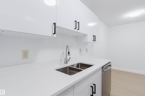 White cabinetry with matte black hardware, dual stainless steel sink, stainless steel dishwasher, and light wood-finish flooring - #106 10520 80 Av Nw, Edmonton, AB - Indoor Photo Showing Kitchen With Double Sink