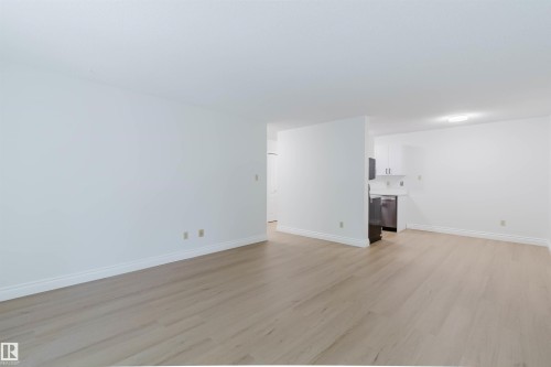 Open-concept living area featuring wood-finish flooring, white walls, and a partial view of the kitchen with stainless steel appliances - #106 10520 80 Av Nw, Edmonton, AB - Indoor Photo Showing Other Room