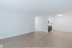 Open-concept living area featuring wood-finish flooring, white walls, and a partial view of the kitchen with stainless steel appliances - 