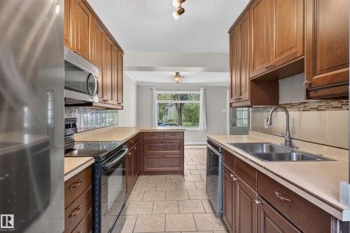 Kitchen featuring wood cabinetry, stainless steel appliances, a double basin sink with a gooseneck faucet, light-toned countertops, and tile flooring - 11549 88 Street Nw, Edmonton, AB - Indoor Photo Showing Kitchen With Double Sink