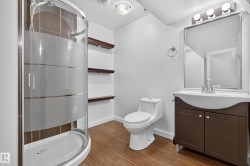 Curved glass shower enclosure with frosted bands, wood-finish flooring, dark wood vanity with integrated basin, framed wall mirror, and built-in wood shelving - 