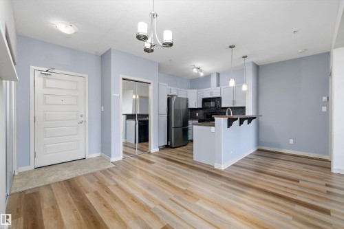 Wood-finish flooring throughout, kitchen with stainless steel appliances, light gray cabinetry, dark countertops, and a breakfast bar with pendant lighting - #207 304 Amblside Li Sw Sw, Edmonton, AB - Indoor Photo Showing Kitchen