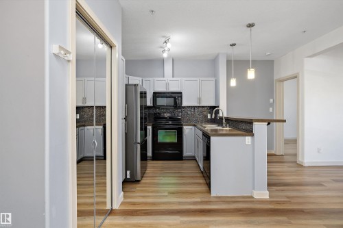 Kitchen with light-toned cabinetry, dark-finish appliances, and a mosaic tile backsplash - #207 304 Amblside Li Sw Sw, Edmonton, AB - Indoor Photo Showing Kitchen With Double Sink With Upgraded Kitchen