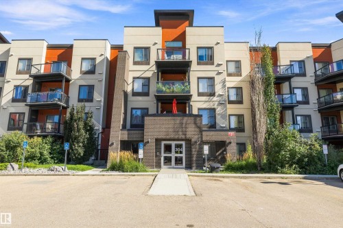 Multi-story building featuring a brick facade, glass balconies, and a paved parking area - #207 304 Amblside Li Sw Sw, Edmonton, AB - Outdoor With Facade
