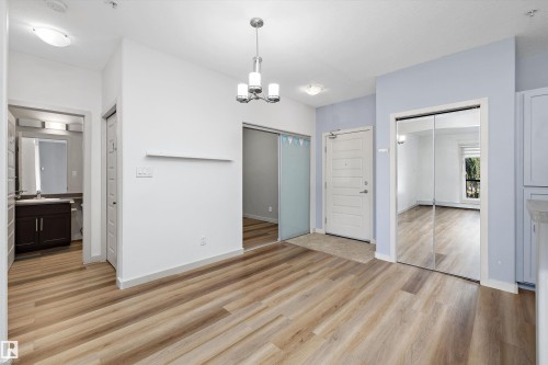 Open-concept living space with wood-finish flooring, a contemporary chandelier, and mirrored closet doors - #207 304 Amblside Li Sw Sw, Edmonton, AB - Indoor Photo Showing Other Room