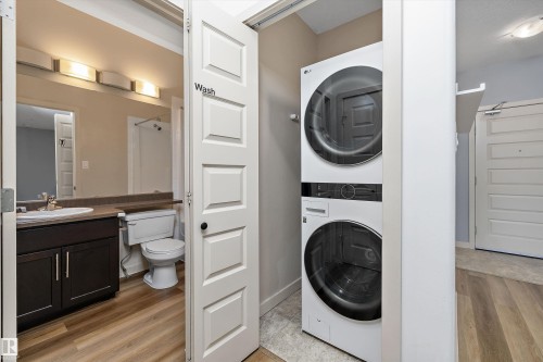 Stacked washer and dryer unit, wood-finish flooring, dark wood-finish vanity with countertop, rectangular sink, and chrome faucet, and a toilet - #207 304 Amblside Li Sw Sw, Edmonton, AB - Indoor Photo Showing Laundry Room