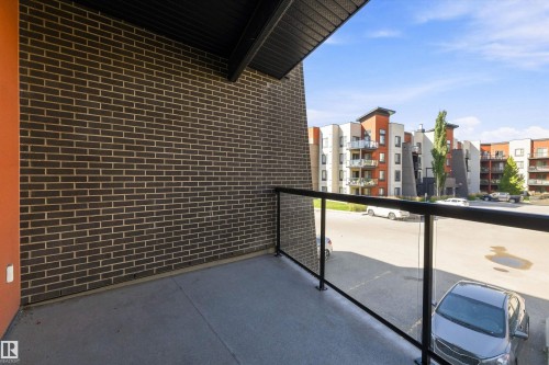 Balcony with concrete flooring and a glass railing, featuring a dark brick exterior wall - #207 304 Amblside Li Sw Sw, Edmonton, AB - Outdoor