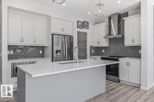 Modern kitchen featuring white cabinetry, stainless steel appliances, a grey subway tile backsplash, a large island with a composite countertop, and wood-finish flooring - 1014 Daniels Loop Sw, Edmonton, AB - Indoor Photo Showing Kitchen With Upgraded Kitchen