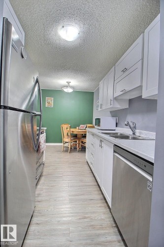 Galley kitchen featuring stainless steel appliances, white cabinetry, light countertops, and wood-finish flooring - #304 14004 26 St Nw, Edmonton, AB - Indoor Photo Showing Kitchen With Double Sink
