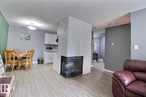 Central fireplace with a black firebox, light gray walls, wood-finish flooring, and white kitchen cabinetry - #304 14004 26 St Nw, Edmonton, AB - Indoor Photo Showing Living Room With Fireplace