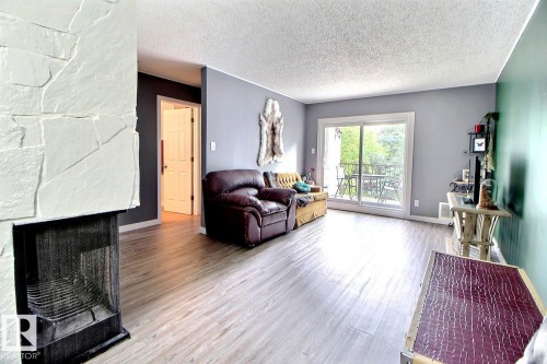 Light wood-finish flooring throughout, a stone-faced fireplace, and sliding glass doors leading to a balcony - #304 14004 26 St Nw, Edmonton, AB - Indoor Photo Showing Living Room