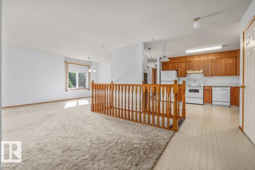 Open-concept living area with neutral carpeting, natural wood banister, and a kitchen featuring wood cabinetry and white appliances, including a range and refrigerator - 4427 Payne Avenue, Onoway, AB - Indoor Photo Showing Kitchen