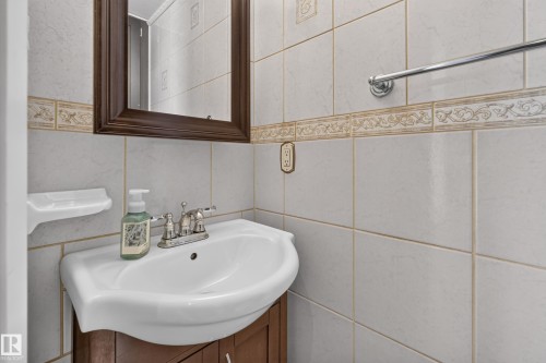 Wall-mounted mirror with a wood-finish frame, ceramic wall tiling with decorative border, white ceramic basin with a dual-handle faucet, and chrome towel bar - 23 Willowdale Place Nw, Edmonton, AB 