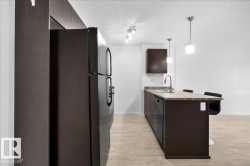 Kitchen featuring dark-finish cabinetry, light-tone wood-finish flooring, a contemporary island with seating, a built-in dishwasher, and pendant lighting - 