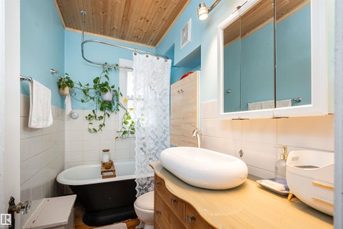 Wood-paneled ceiling, light blue walls, white tiled lower walls, freestanding black bathtub, and wood-finish vanity with an oval vessel sink - 11506 94 Street Nw, Edmonton, AB 