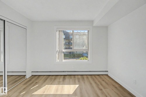 Wood-finish flooring, white walls, and a window with white trim, featuring a mirrored sliding closet door - #226 17011 67 Av Nw, Edmonton, AB - Indoor Photo Showing Other Room