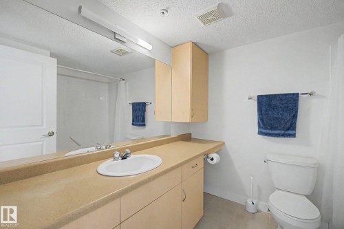 Integrated vanity with an oval sink and chrome faucet, full-width mirror, wall-mounted cabinetry, and a toilet - #226 17011 67 Av Nw, Edmonton, AB - Indoor Photo Showing Bathroom