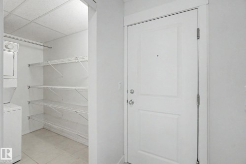 White paneled door with polished chrome hardware, adjacent to a utility closet featuring a stacked washer and dryer, wire shelving, and a hanging rod - #226 17011 67 Av Nw, Edmonton, AB - Indoor With Storage