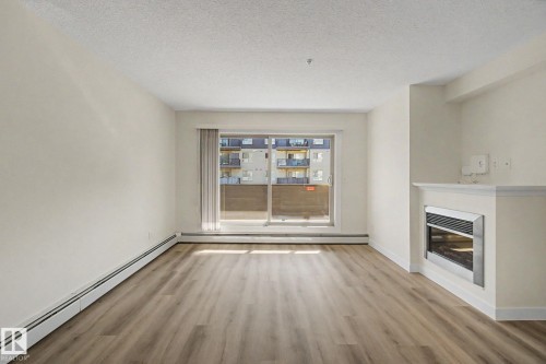 Spacious room featuring wood-finish flooring, a built-in fireplace with a mantel, and a sliding glass door - #226 17011 67 Av Nw, Edmonton, AB - Indoor With Fireplace