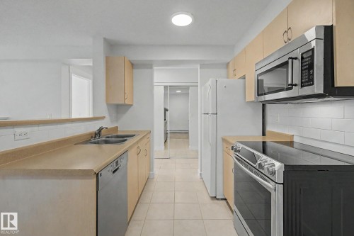 Galley kitchen with light wood-finish cabinetry, tile flooring, and integrated stainless steel appliances including an oven and microwave - #226 17011 67 Av Nw, Edmonton, AB - Indoor Photo Showing Kitchen With Double Sink