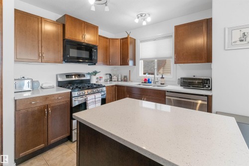 Kitchen featuring wood-finish cabinetry, stainless steel appliances, a center island with a light-toned countertop, and tiled flooring - 6515 37 Avenue, Beaumont, AB - Indoor Photo Showing Kitchen With Double Sink