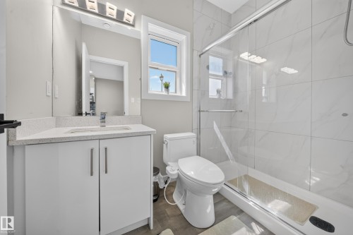 Modern bathroom featuring a white vanity with dual handles and an integrated sink, quartz countertop, large framed mirror with integrated lighting, walk-in shower with glass enclosure and tile surround, and a ceramic toilet with elongated bowl - 4301 38 St, Beaumont, AB - Indoor Photo Showing Bathroom