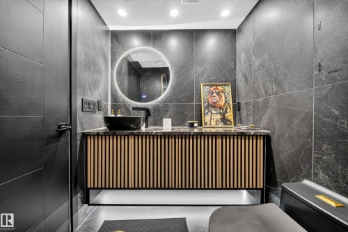 Full-height dark stone tile walls, a circular backlit mirror, a vessel sink with a matte black faucet, and a contemporary vanity with a slatted wood-finish front - 4301 38 St, Beaumont, AB - Indoor Photo Showing Other Room