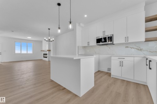 White cabinetry with matte black hardware, integrated microwave, wood-finish flooring, white island, and fireplace with white brick surround - 82 Patriot Way, Spruce Grove, AB - Indoor Photo Showing Kitchen