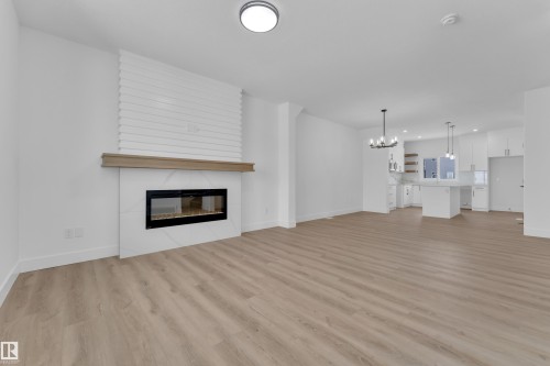 Expansive wood-finish flooring, contemporary linear fireplace with stone surround, white brick-patterned fireplace facade, modern kitchen with white cabinetry, and a central island with pendant lighting - 82 Patriot Way, Spruce Grove, AB - Indoor Photo Showing Living Room With Fireplace