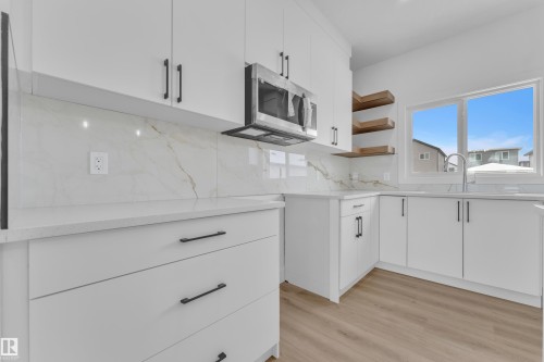 White cabinetry with matte black hardware, integrated stainless steel microwave, light-toned wood-finish flooring, white countertops, and a white tile backsplash - 82 Patriot Way, Spruce Grove, AB - Indoor Photo Showing Kitchen