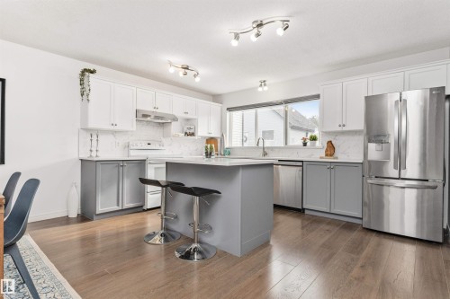 Modern kitchen featuring two-tone cabinetry, stainless steel appliances, a center island with quartz countertop seating, Vinyl plank flooring, and track lighting - 4121 Tompkins Way Nw, Edmonton, AB - Indoor Photo Showing Kitchen With Upgraded Kitchen