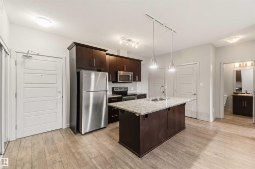 Kitchen featuring dark wood cabinetry, stainless steel appliances, a center island with a granite countertop, and pendant lighting - #414 5510 Schonsee Dr Nw, Edmonton, AB - Indoor Photo Showing Kitchen With Stainless Steel Kitchen With Double Sink