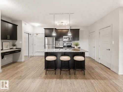 Open-concept kitchen with dark wood-finish cabinetry, stainless steel appliances, a center island with countertop seating, and light wood-finish flooring - #414 5510 Schonsee Dr Nw, Edmonton, AB - Indoor