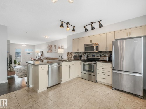Open-concept kitchen featuring light-toned cabinetry, stainless steel appliances, a breakfast bar, stone countertops, and a mosaic tile backsplash - #21 1140 Chappelle Bv Sw, Edmonton, AB - Indoor Photo Showing Kitchen