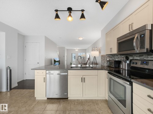Kitchen featuring beige cabinetry, stainless steel appliances, dark countertops, and a mosaic tile backsplash - #21 1140 Chappelle Bv Sw, Edmonton, AB - Indoor Photo Showing Kitchen