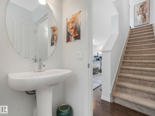 Pedestal sink with chrome faucet and oval mirror, complemented by wood-finish flooring and a carpeted staircase - #21 1140 Chappelle Bv Sw, Edmonton, AB - Indoor Photo Showing Bathroom