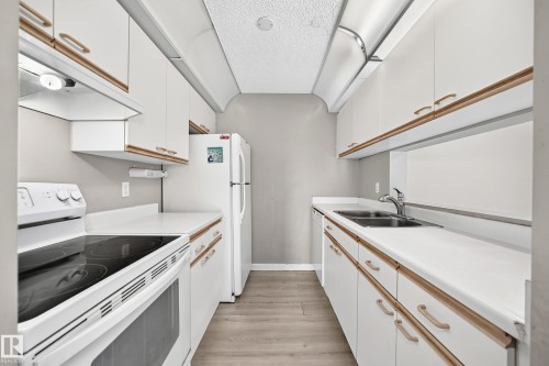Galley kitchen featuring wood-finish flooring, white cabinetry with wood trim, white countertops, a double basin stainless steel sink, and a white electric range - #204 11218 80 St Nw, Edmonton, AB - Indoor Photo Showing Kitchen With Double Sink
