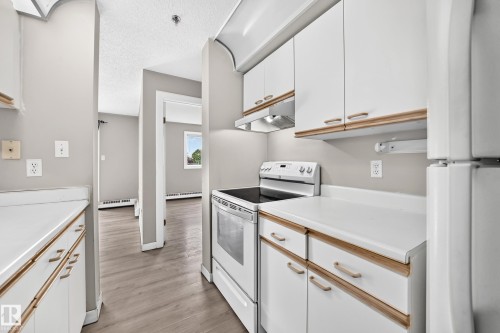 Galley kitchen with white cabinetry and wood-finish trim, white solid surface countertops, and a white electric range - #204 11218 80 St Nw, Edmonton, AB - Indoor Photo Showing Kitchen