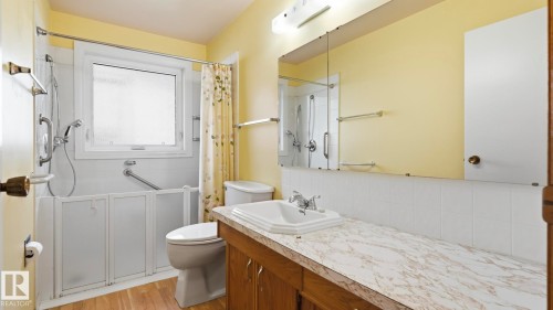 Shower-tub combination with grab bars, wood-finish flooring, vanity with wood-finish cabinetry, countertop with integrated sink, and large wall-mounted mirror - 8408 176 Street Nw, Edmonton, AB - Indoor Photo Showing Bathroom