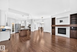 Open-concept design featuring wood-finish flooring, a kitchen island with pendant lighting, integrated stainless steel appliances, a built-in media center with shelving, and a gas fireplace - 