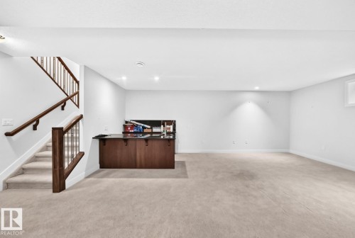 Spacious room featuring light carpeting, a built-in wet bar with dark cabinetry, and a staircase with wood railings - #57 18343 Lessard Rd Nw, Edmonton, AB - Indoor Photo Showing Other Room