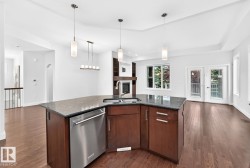 Spacious open-concept living area featuring wood-finish flooring, a kitchen island with dark cabinetry and a stainless steel dishwasher, a built-in fireplace, and double French doors to an exterior space - 