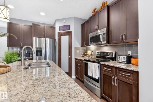 Spacious kitchen featuring granite-look countertops, dark wood-finish cabinetry, a large stainless steel sink with a gooseneck faucet, and a subway tile backsplash - 16512 140 Street Nw, Edmonton, AB - Indoor Photo Showing Kitchen With Double Sink With Upgraded Kitchen